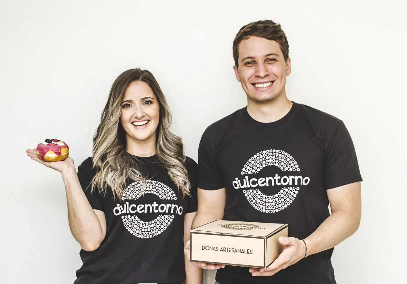 Andreina y José María, fundadores de Dulcentorno, con poleras de la marca, una dona y una caja de donas artesanales