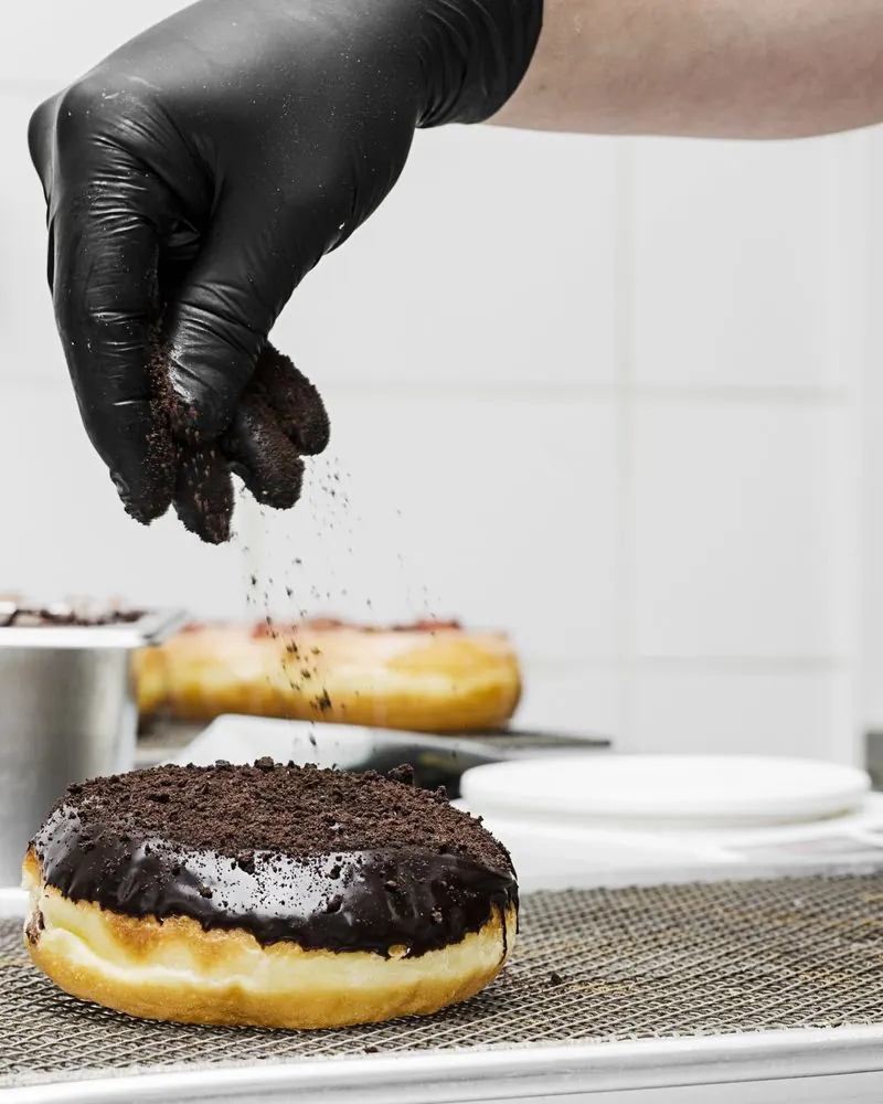 Mano con guante espolvoreando galleta triturada sobre una dona bañada en chocolate
